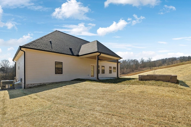 rear view of house featuring a lawn, cooling unit, a shingled roof, and a patio