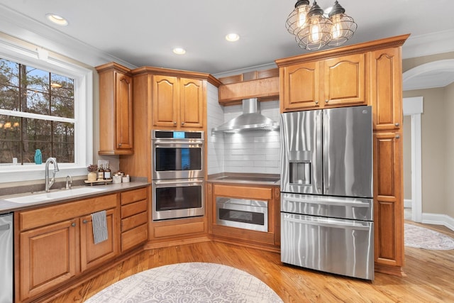 kitchen with light wood finished floors, arched walkways, a sink, stainless steel appliances, and wall chimney exhaust hood