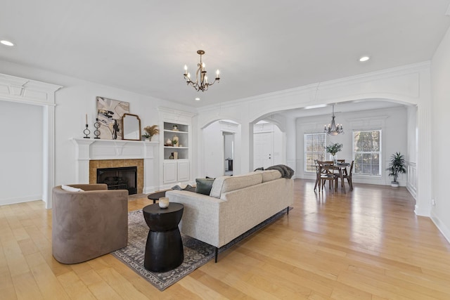 living room featuring built in features, light wood-type flooring, a tile fireplace, arched walkways, and a notable chandelier