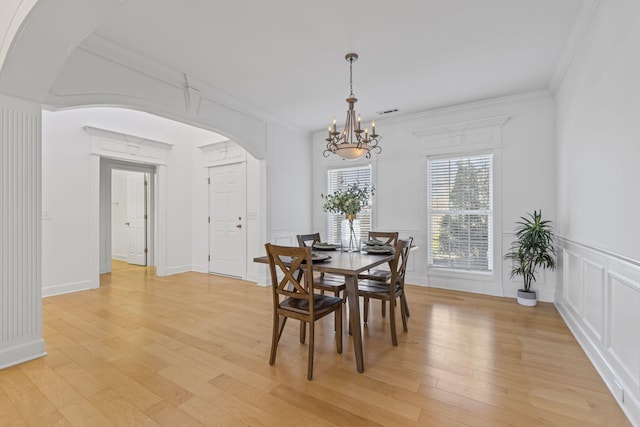 dining area with light wood-style flooring, arched walkways, ornamental molding, a decorative wall, and a notable chandelier