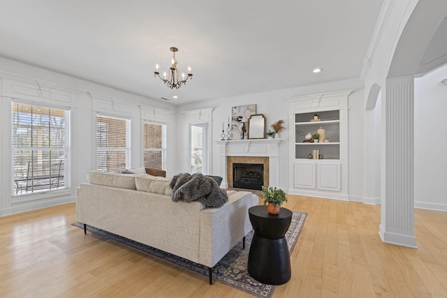 living room with light wood-type flooring, a tiled fireplace, built in features, an inviting chandelier, and ornate columns