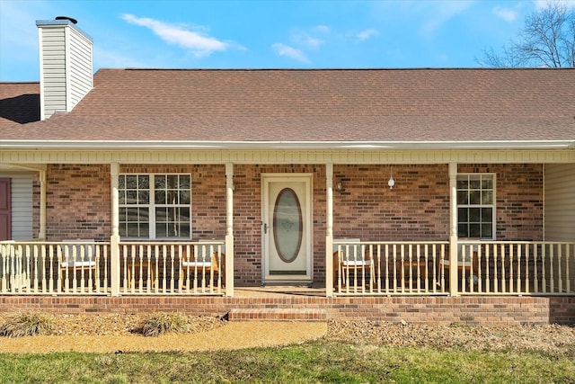 doorway to property featuring brick siding, covered porch, a chimney, and roof with shingles