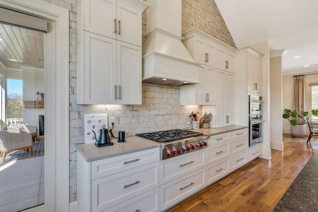 kitchen featuring light wood-style flooring, ornamental molding, custom range hood, backsplash, and appliances with stainless steel finishes