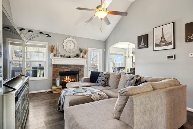 living room featuring dark wood-style floors, baseboards, arched walkways, a stone fireplace, and ceiling fan with notable chandelier