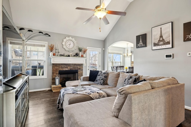 living area with ceiling fan with notable chandelier, dark wood finished floors, arched walkways, a stone fireplace, and baseboards