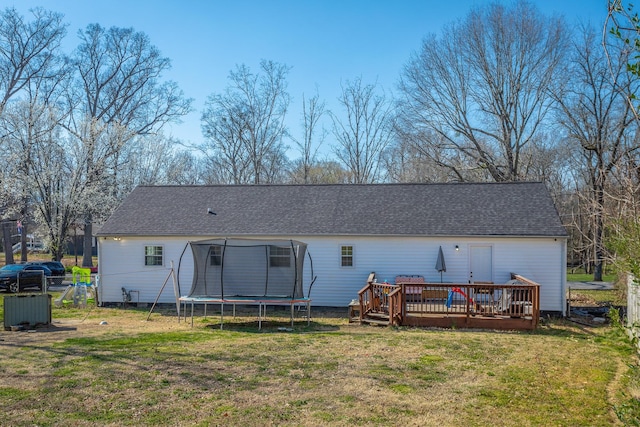 rear view of property featuring a trampoline, fence, roof with shingles, a lawn, and a deck