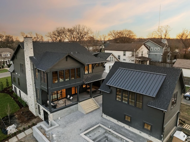 back of house at dusk featuring a standing seam roof, a residential view, roof with shingles, a chimney, and a patio area