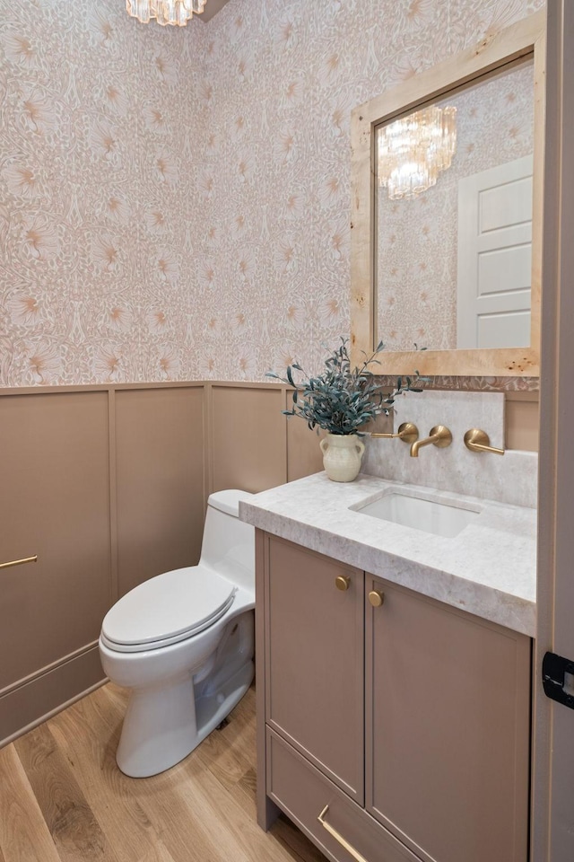 half bathroom featuring wallpapered walls, toilet, wainscoting, a notable chandelier, and vanity