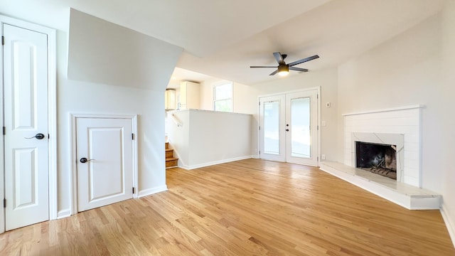 unfurnished living room featuring light wood-type flooring, a fireplace with raised hearth, a ceiling fan, french doors, and baseboards