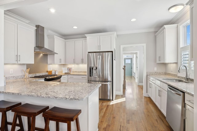 kitchen featuring light wood finished floors, a peninsula, a sink, stainless steel appliances, and wall chimney range hood