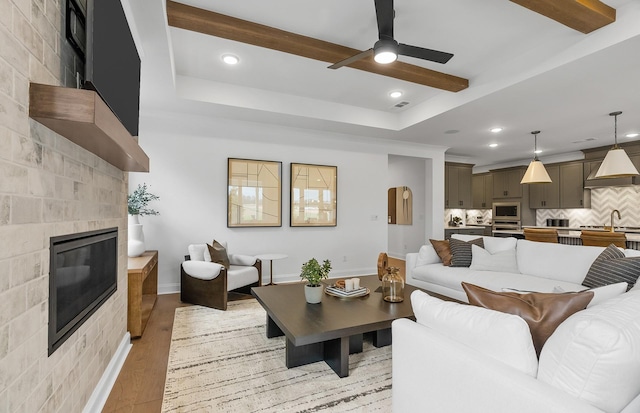 living area with baseboards, beamed ceiling, light wood-style floors, a glass covered fireplace, and a ceiling fan