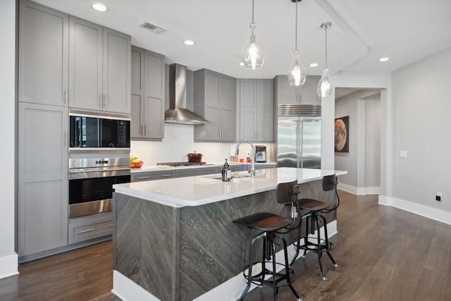 kitchen with a sink, built in appliances, gray cabinetry, and wall chimney range hood