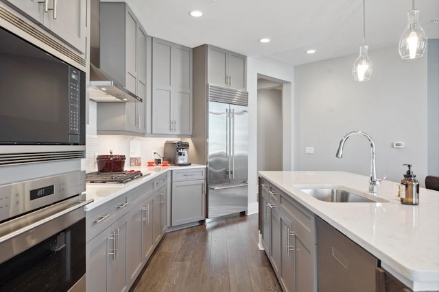 kitchen with gray cabinetry, a sink, dark wood finished floors, wall chimney range hood, and built in appliances