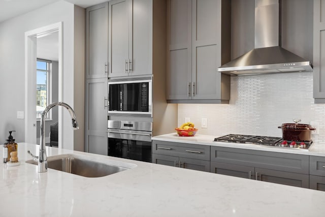 kitchen featuring gray cabinetry, a sink, wall chimney exhaust hood, light stone countertops, and stainless steel gas cooktop