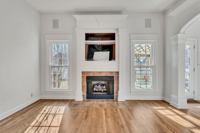 unfurnished living room featuring a tiled fireplace, ornate columns, a wealth of natural light, and wood finished floors