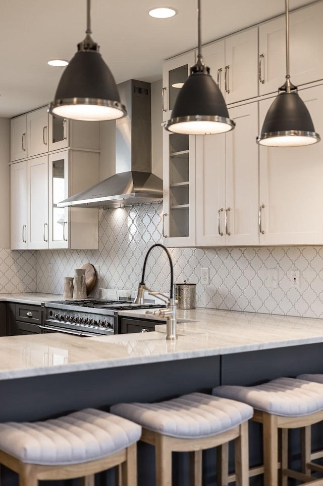 kitchen with a breakfast bar area, light stone countertops, glass insert cabinets, and wall chimney exhaust hood