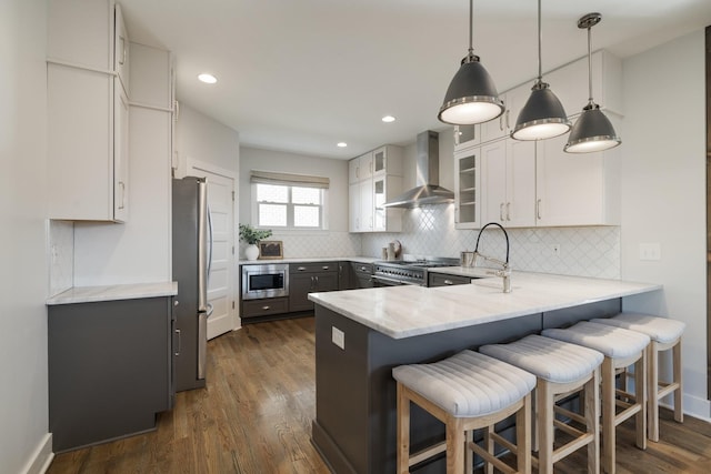 kitchen with backsplash, a peninsula, appliances with stainless steel finishes, wall chimney exhaust hood, and light stone countertops