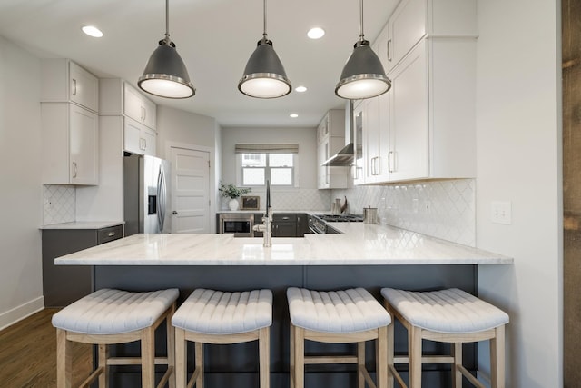 kitchen with a sink, stainless steel appliances, a peninsula, wall chimney range hood, and light stone countertops