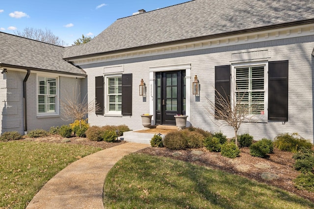 view of exterior entry with brick siding, a shingled roof, and a yard