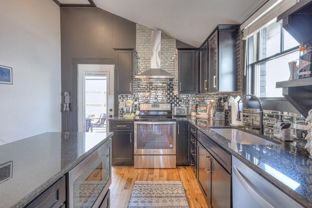 kitchen with light wood finished floors, vaulted ceiling, appliances with stainless steel finishes, wall chimney exhaust hood, and a sink