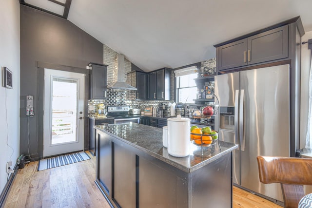 kitchen with dark stone countertops, light wood finished floors, stainless steel appliances, vaulted ceiling, and wall chimney exhaust hood