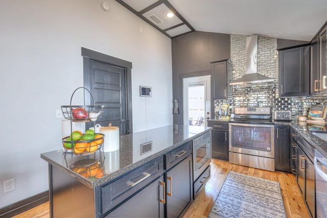 kitchen featuring light wood finished floors, wall chimney range hood, vaulted ceiling, dark stone countertops, and stainless steel appliances