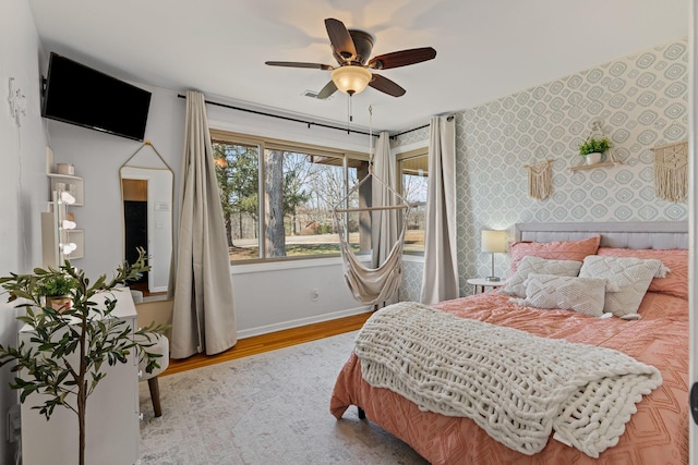 bedroom with wallpapered walls, an accent wall, baseboards, wood finished floors, and a ceiling fan