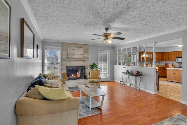 living room featuring a ceiling fan, light wood finished floors, a textured ceiling, crown molding, and a brick fireplace