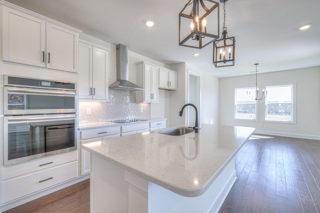 kitchen with a sink, a chandelier, stainless steel double oven, wall chimney exhaust hood, and a kitchen island with sink