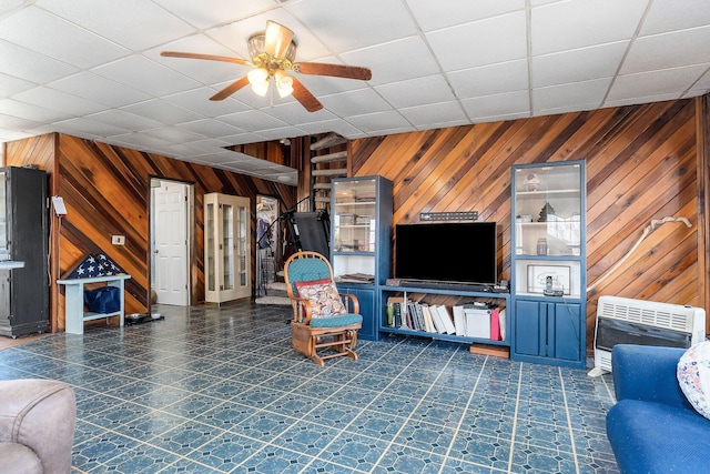 unfurnished living room with a drop ceiling, heating unit, wood walls, and ceiling fan