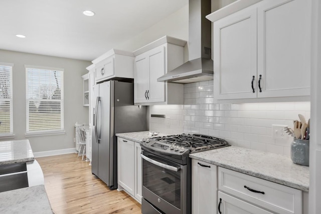 kitchen featuring light wood-style flooring, stainless steel appliances, white cabinetry, wall chimney range hood, and backsplash