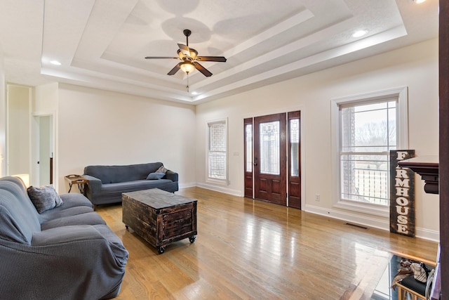 living room with a ceiling fan, visible vents, light wood finished floors, baseboards, and a tray ceiling