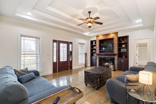 living room with light wood-type flooring, a ceiling fan, a tray ceiling, a large fireplace, and baseboards