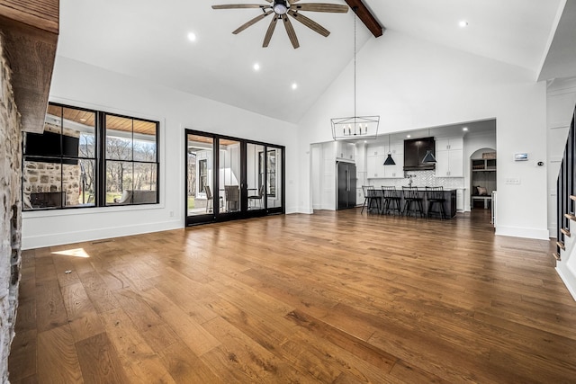 unfurnished living room featuring stairway, dark wood-style floors, high vaulted ceiling, beamed ceiling, and ceiling fan with notable chandelier