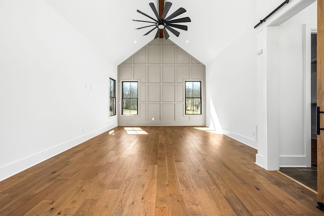 unfurnished living room with baseboards, ceiling fan, a barn door, hardwood / wood-style flooring, and high vaulted ceiling