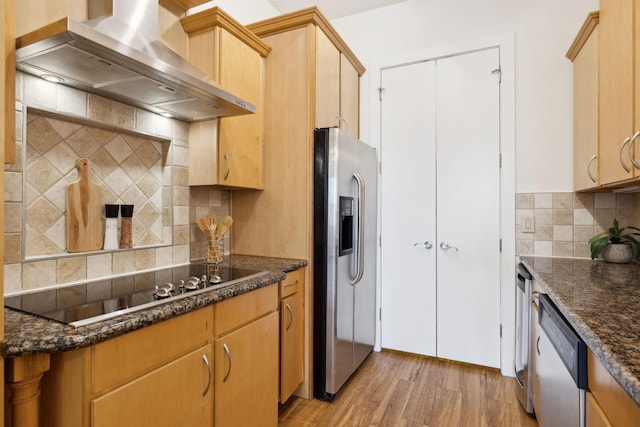 kitchen featuring wall chimney range hood, light wood-type flooring, decorative backsplash, dark stone countertops, and appliances with stainless steel finishes