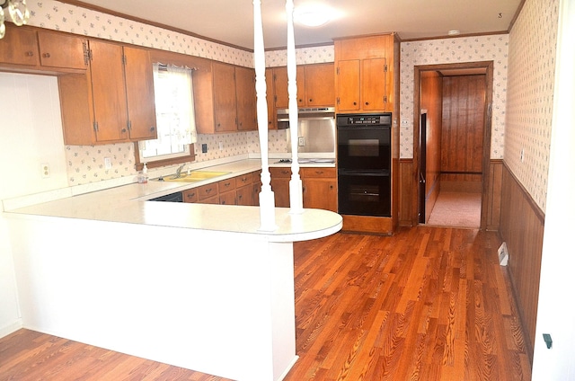 kitchen with a sink, wallpapered walls, a peninsula, wainscoting, and light countertops