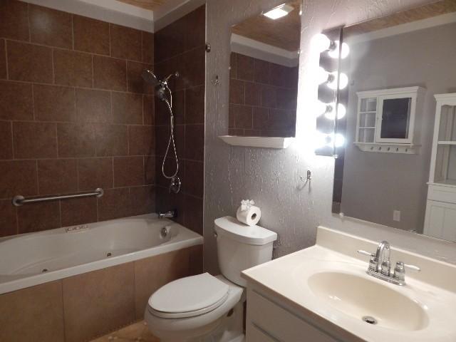 full bath with vanity, toilet, a textured wall, and a combined bath / shower with jetted tub