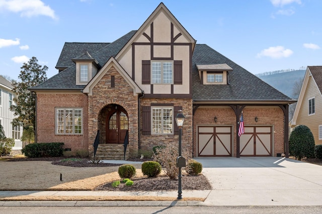 tudor house with brick siding, an attached garage, a shingled roof, stucco siding, and driveway