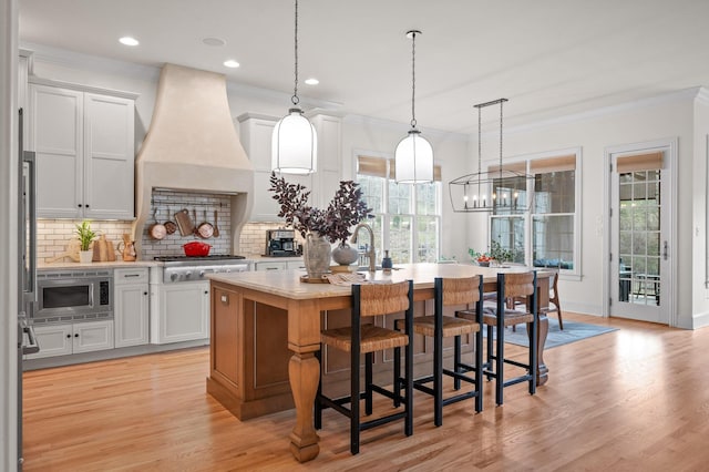 kitchen featuring a center island with sink, ornamental molding, light countertops, custom range hood, and appliances with stainless steel finishes