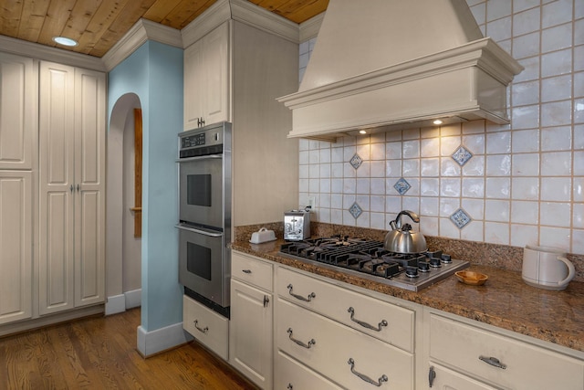 kitchen with decorative backsplash, arched walkways, wood finished floors, custom exhaust hood, and stainless steel appliances