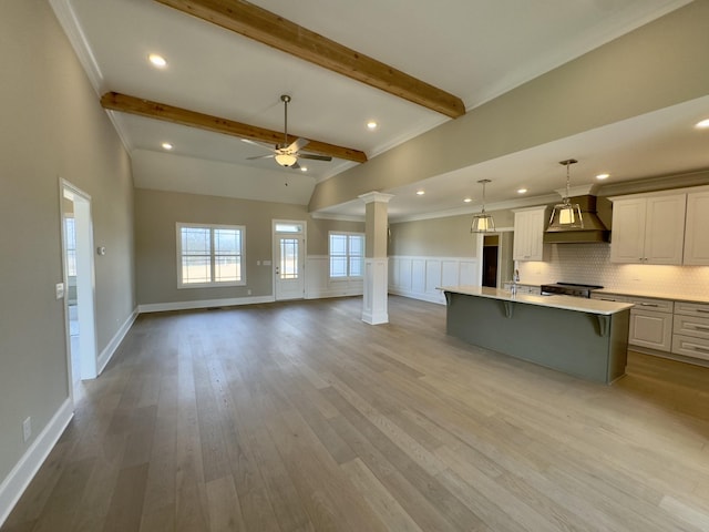 kitchen with light wood-type flooring, a center island with sink, light countertops, decorative columns, and custom exhaust hood