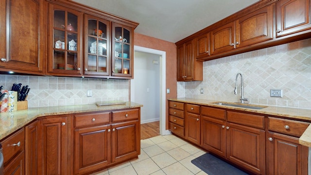 kitchen featuring brown cabinets, a sink, light stone counters, light tile patterned flooring, and glass insert cabinets