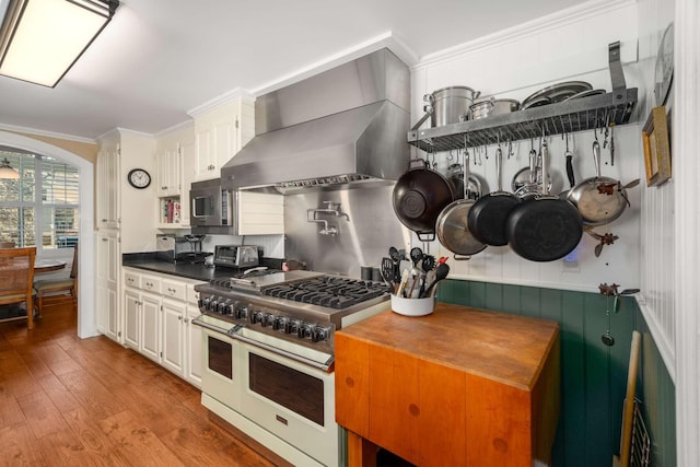 kitchen with ornamental molding, double oven range, stainless steel microwave, wood-type flooring, and wall chimney range hood