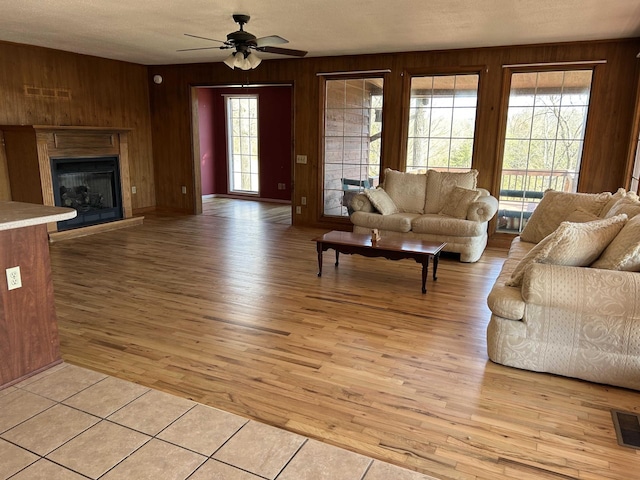 living room featuring light wood-type flooring, visible vents, a glass covered fireplace, wooden walls, and ceiling fan