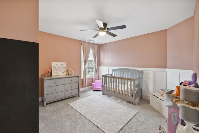 carpeted bedroom featuring visible vents, a ceiling fan, a nursery area, and a wainscoted wall