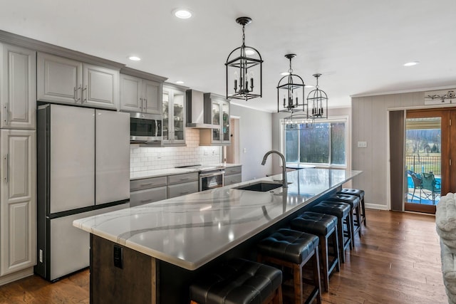 kitchen featuring a center island with sink, gray cabinets, a sink, stainless steel appliances, and wall chimney exhaust hood