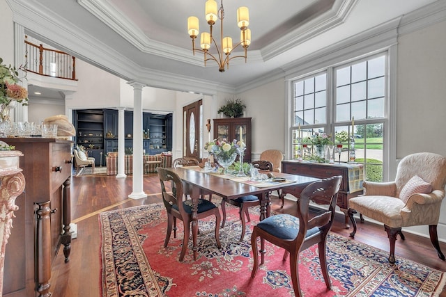 dining room featuring ornamental molding, wood finished floors, a notable chandelier, a raised ceiling, and ornate columns