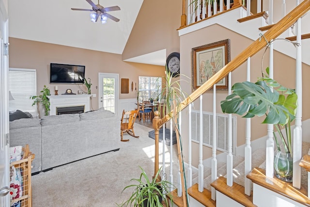 carpeted living room featuring ceiling fan, stairs, wainscoting, a fireplace, and high vaulted ceiling