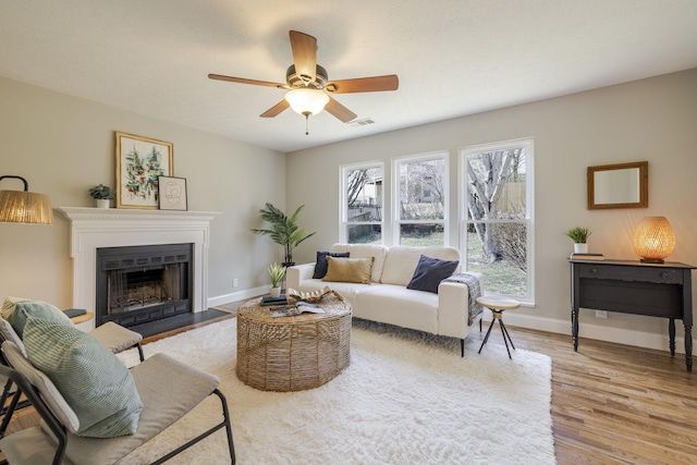 living area featuring baseboards, a fireplace with flush hearth, light wood-style flooring, and a ceiling fan